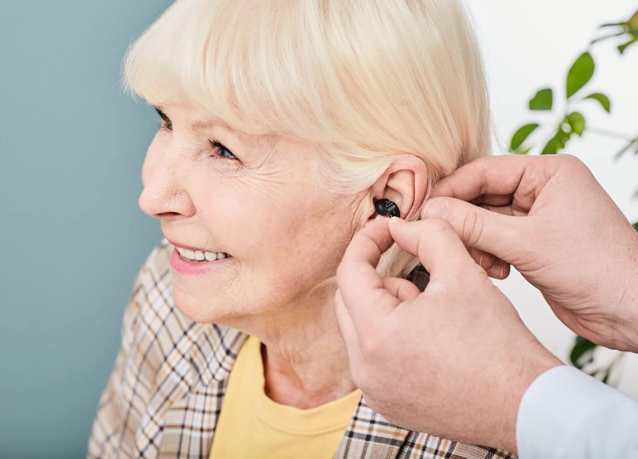 Audiologist fitting a cheerful senior woman with a hearing aid.