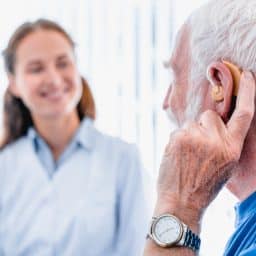 an elderly male patient with hearing aid is pictured with an audiologist in the background